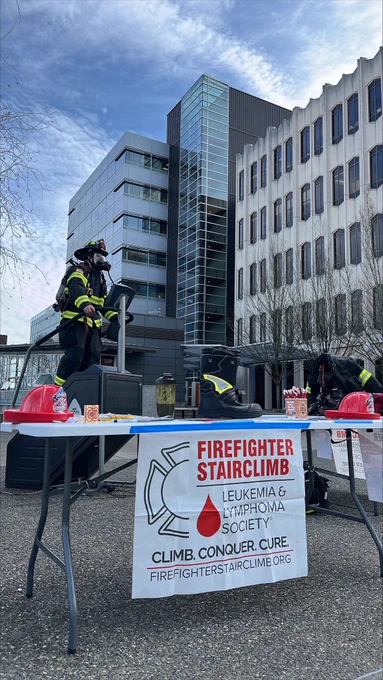 A firefighter in full turnout gear, with breathing apparatus and air tank, climbs on a stair climber in front of Snohomish County government buildings. A folding table is set up in front of the stair climber with red plastic firefighter hats and a sign that reads Firefighter Stairclimb with the official Leukemia & Lymphoma Society logo.