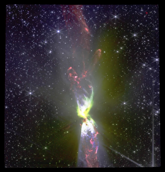 A NIRCam (Near-Infrared Camera) image of the dark cloud Lynds 483, highlighting a powerful bipolar jet. The jet is brightly illuminated with a mix of red, green, and blue hues, indicating different ionized gases and dust. It extends vertically from a bright, star-forming region at the bottom, against a backdrop of numerous faint stars. The image showcases the detailed structure and complex emissions within the cloud.