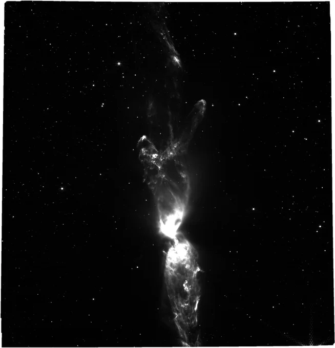 A raw, black and white image of Lynds 483 showcasing a prominent, vertical jet structure emanating from a bright, compact source at the bottom. The jet appears as a luminous, textured column against a stark black background punctuated by numerous faint stars. The image emphasizes the structural details of the outflow without color information.