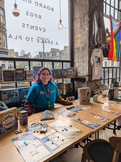 Board games set up for demos in the board game shop Ancient Robot Games. Asmodee demonstrator is smiling by the table of games.