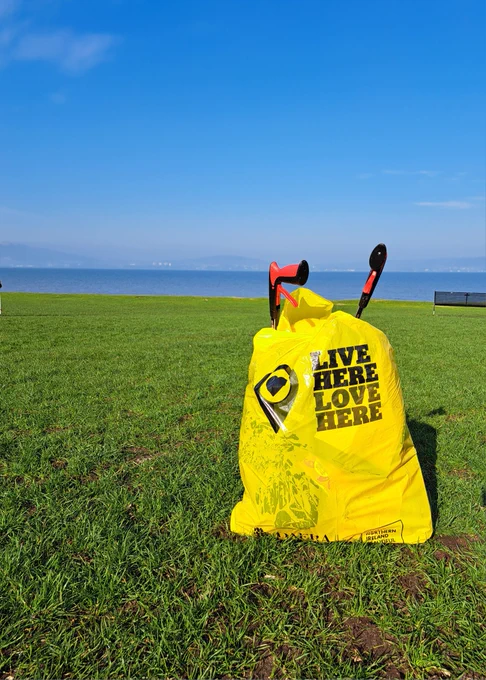 A photo of a full bag of litter collected on the parkland with 2 litterpickers, the sea is pictured in the background 