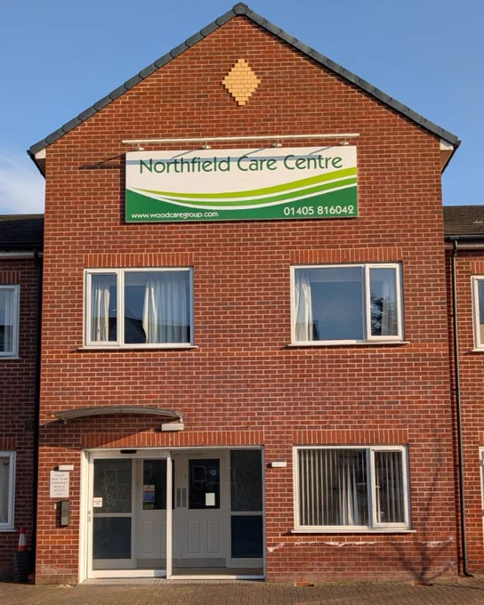 Front elevation of a building featuring a large white sign with green graphics applied.  The top of the sign has the text 'Northfield Care Centre' and the bottom edge has a green swooping design across it.