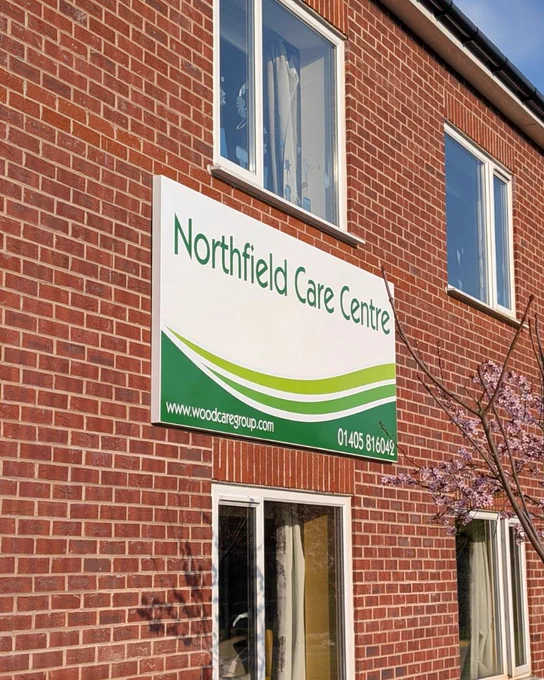 A building featuring a white sign with green graphics applied.  The top of the sign has the text 'Northfield Care Centre' and the bottom edge has a green swooping design across it.  A blossom tree is adjacent to the sign.