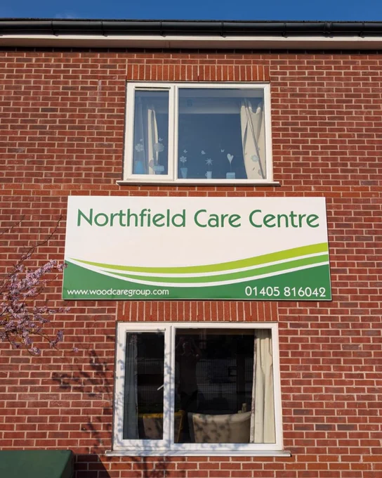 A red brick building featuring a white sign with green graphics applied.  The top of the sign has the text 'Northfield Care Centre' and the bottom edge has a green swooping design across it.  A blossom tree encroaches onto the left side of the sign.