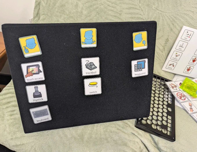 The image shows a black board with various printed symbols attached to it. The symbols are arranged in a grid pattern and show different assistive technology tools. The top row has three icons with a blue character showing a thumbs up, a character with arms outstretched, and a character with a thumbs down. In the column beneath the character showing thumbs up, there are symbols. From top to bottom they read "touch access" with a hand pointing to a touchscreen, "joystick" with an image of a joystick and "keyboard" with an image of a keyboard. The second column beneath the character with arms outstretched are two symbols, "trackball" with an image of a trackball device and "switch" with an image of a yellow switch button. In the last column beneath the character with thumbs down is a symbol with a "keyguard" with an image of a keyguard for a keyboard.