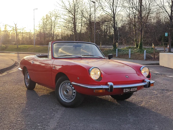 A Fiat 850 Spider, in red, front three quarter view. Photographed into the low sun