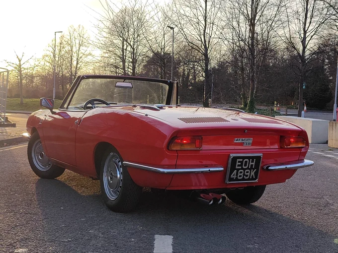 A Fiat 850 Spider, in red, rear three quarter view. Photographed into the low sun 