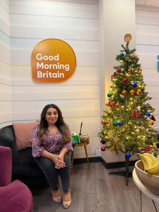 Shani, a South Asian disabled woman sitting on a blue chair with the Good Morning Britain logo behind her on the wall. She’s sitting in the green room where the show is filmed and there is a lit up Christmas tree next to her and a bowl of bananas on the table.