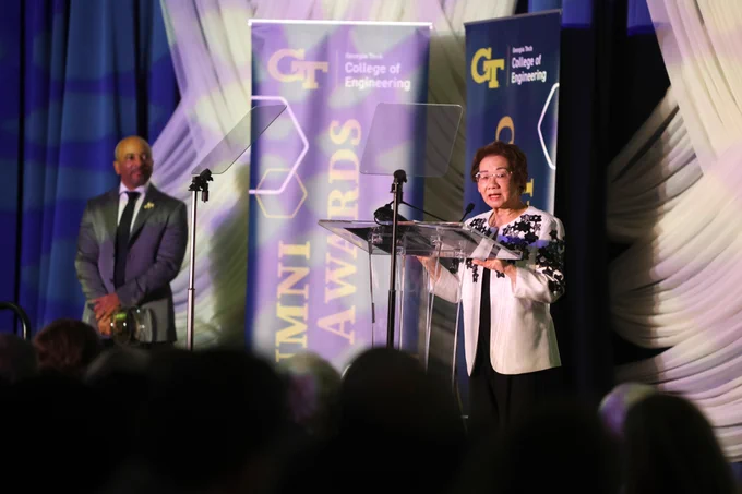 Coulter Foundation President Sue Van speaking at the podium while Dean Raheem Beyah looks on from the side of the stage at the College of Engineering Alumni Awards and Induction Ceremony.