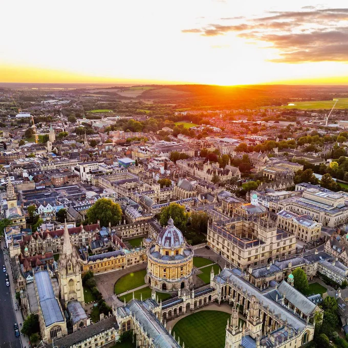 Aerial view of Oxford at sunset, showcasing the historic architecture including the Radcliffe Camera and surrounding university buildings, with a vibrant sky and expansive green landscapes in the background.