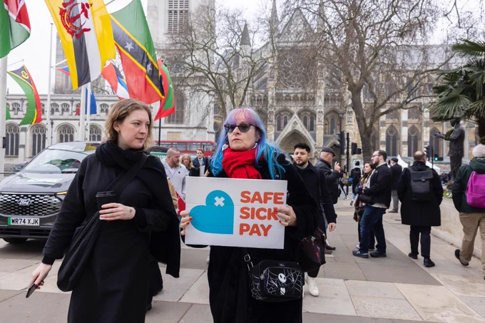 Two women walk through Parliament square holding the Safe Sick Pay placard. Credit: Sylvie Pope Photography, Safe Sick Pay campaign