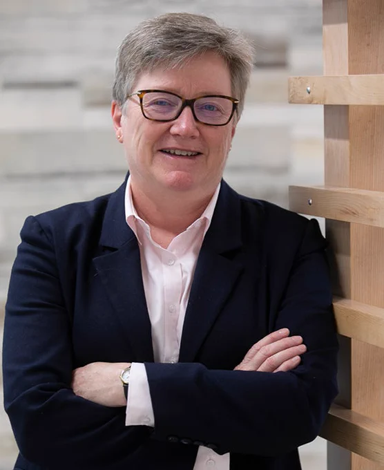 Catherine Clifford smiling confidently with crossed arms, standing next to a wooden structure against a blurred, light-colored background. She are dressed in a dark blazer over a light pink collared shirt.