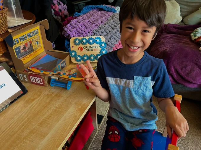 A child with a big smile sits at a small desk with a CrunchLabs Build Box on it. He has a wooden diploma with eleven gears and an empty spot. 