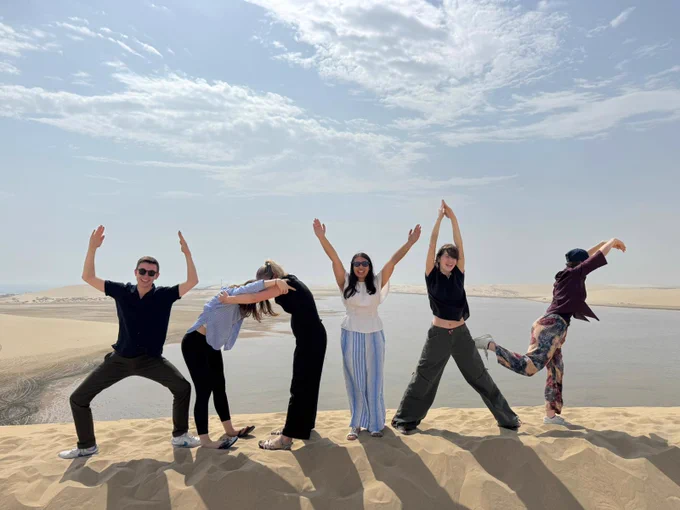 Six students posing playfully to spell "HOYAS" on a sand dune under a clear sky.