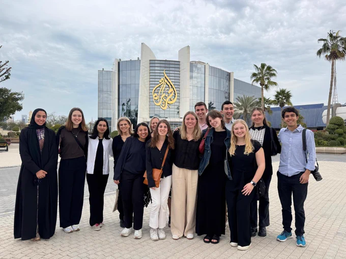 Group of people smiling in front of the Al Jazeera headquarters in Doha, with palm trees and a modern building in the background.
