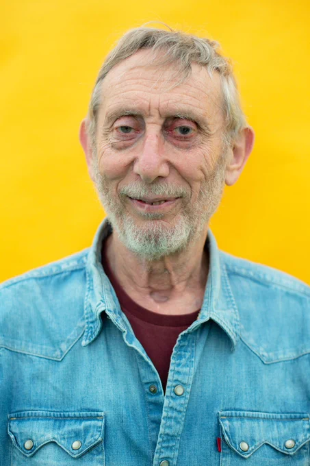 A portrait of Michael Rosen, he is smiling and standing behind a vibrant yellow wall as he wears a relaxed open collar denim shirt.