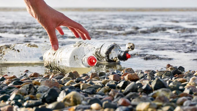 A hand reaches for plastic bottles on a rocky beach, with the ocean in the background