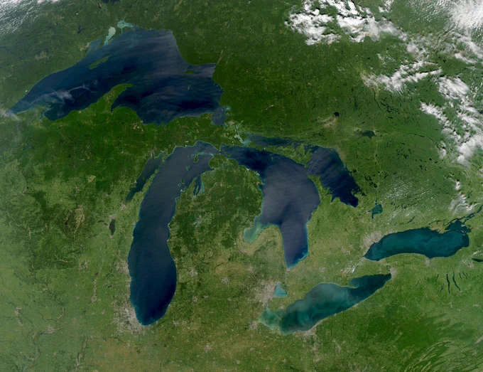 The photo is a satellite image of the Great Lakes region in North America. It prominently features the five Great Lakes—Superior, Michigan, Huron, Erie, and Ontario—surrounded by lush green landmasses, including parts of the United States and Canada. The image shows varying shades of blue in the lakes, with some cloud cover scattered across the land.
