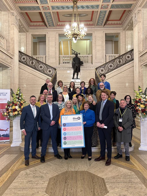 25 people stand on the steps in Stormont Great Hall, two at the front holding a ‘Give 5’ poster.
