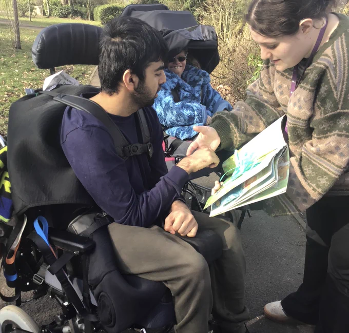 A young man in a powerchair is taking part in a sensory story. A Sense support worker standing in front of him and guides his hand to touch the book that she is holding. Behind him is a young woman in a powerchair. 