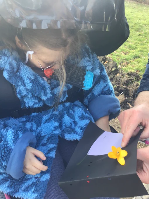 A young woman wearing a blue fluffy fleece is exploring a yellow flower in an outdoor space. 