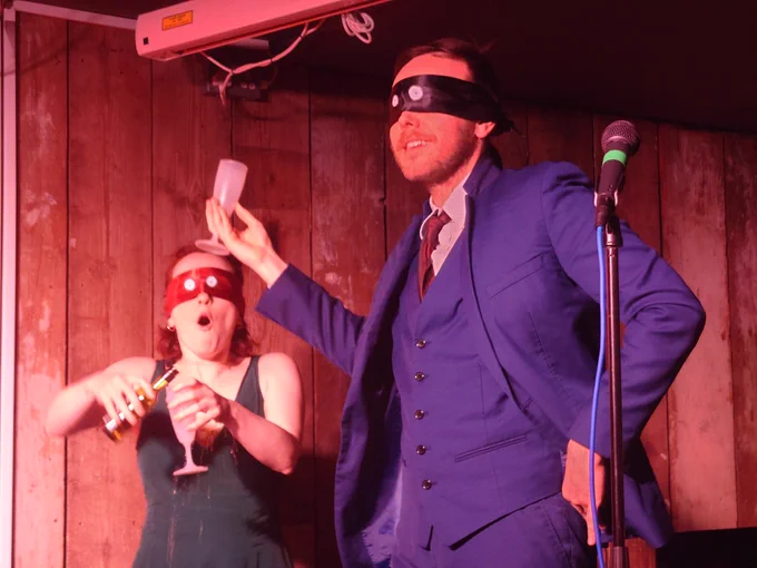 Two blindfolded people in fancy outfits try to pour wine