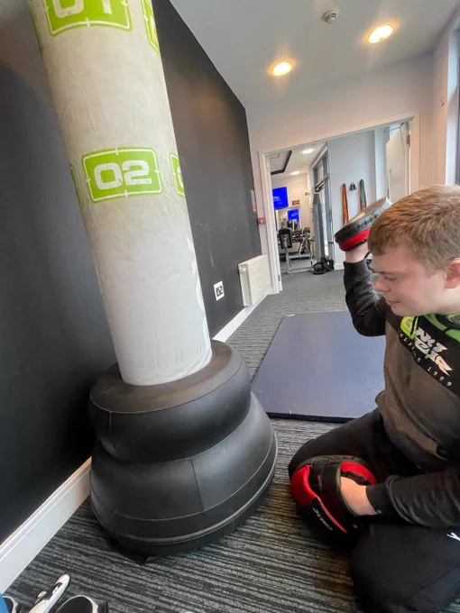 A young man wearing a tracksuit sitting on the floor in front of a boxing punch bag. He is wearing boxing pads on his hands. 