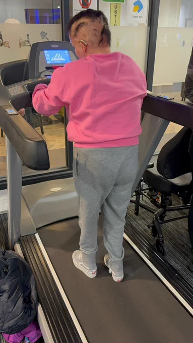 A young woman wearing a hearing aid, pink sweatshirt and grey tracksuit bottoms using a treadmill. 