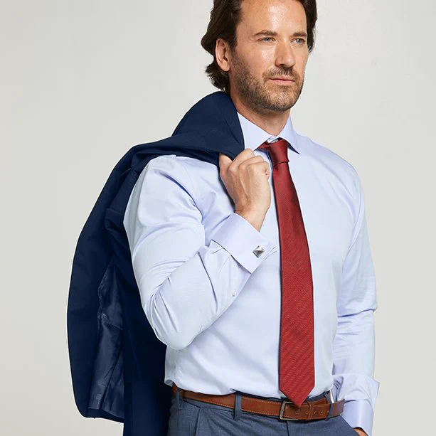 man holding a navy suit jacket over his shoulder,  wearing a blue twill non iron shirt by double two and a red doubletwo tie. he is also wearing navy formal trousers with  a brown belt