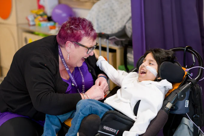Anne and Yusuf are staring at each other, smiling widely. Yusuf is sat in his wheelchair, with his hand on Anne's shoulder, while Anne has her arms in his lap.