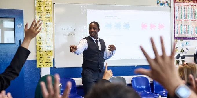 A teacher at the front of the classroom pointing to a student raising their hand, amidst a sea of students raising their hands to answer a question. Credit: Lucy Pope.