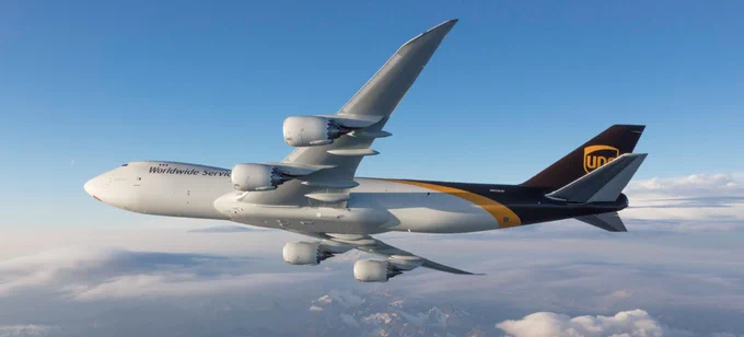 A UPS cargo airplane flies above the clouds. The aircraft has a white fuselage with 'Worldwide Services' written on it and a brown tail featuring the UPS logo. The sky is clear with some scattered clouds below, and the horizon shows distant mountain peaks.