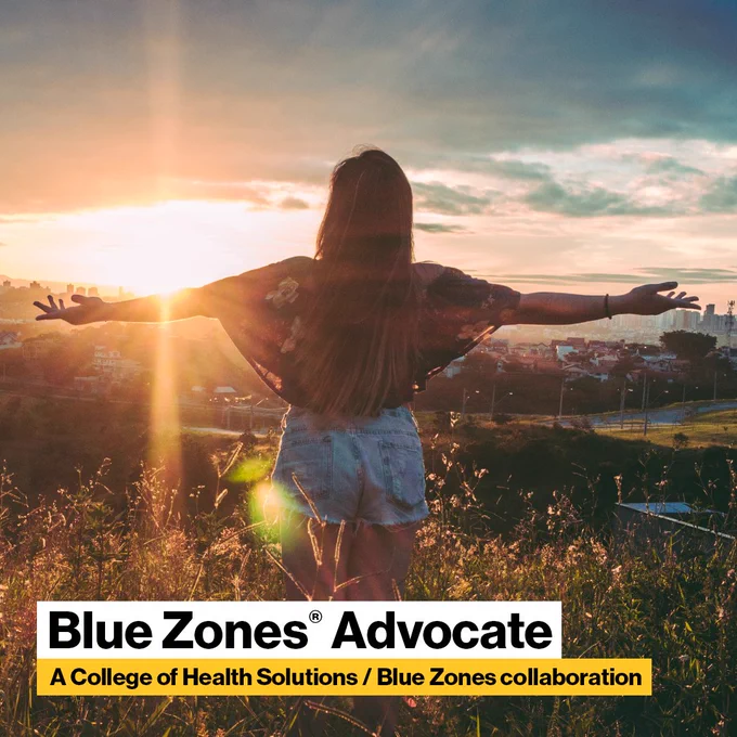 Seen from behind, a woman stretches her arms wide as she stands in a field looking toward the setting sun. At bottom is type that reads, Blue Zones Advocate, a College of Health Solutions / Blue Zones collaboration.