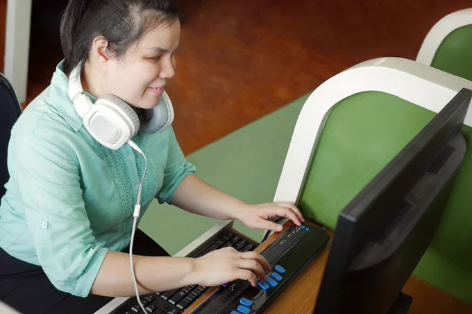 A blind woman wearing headphones using a computer with a braille device.