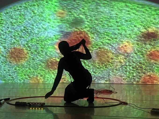 A performer silhouetted by a large projection of green and orange microbes, with small pieces of technology surrounding her on the floor.
