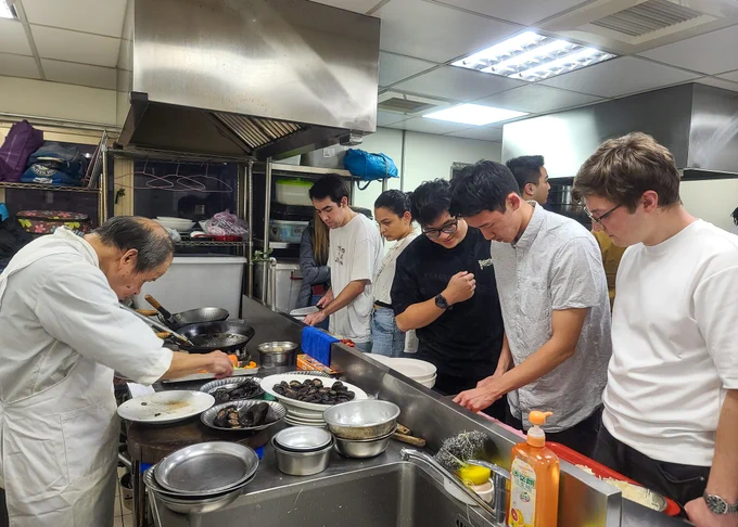 A group of students attentively watch a chef cooking in a commercial kitchen.