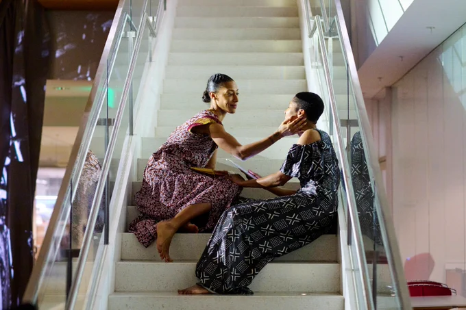 Two women sit on the white steps of a glass-walled staircase. They are looking at each other, as one touches the other's face. 