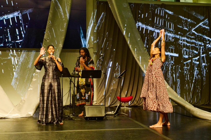 Three performers appear on a stage draped in translucent fabric. 