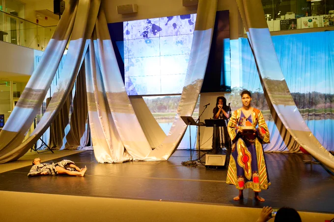 Professor Danielle Wood stands at the front of the stage. Another performer is lying on the ground stage right, while a third plays a musical instrument upstage. Maps and landscape images are projected on the walls behind them. 