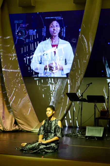Professor Danielle Wood sits at the front of the stage, below a projection of a woman in a white lab coat. 