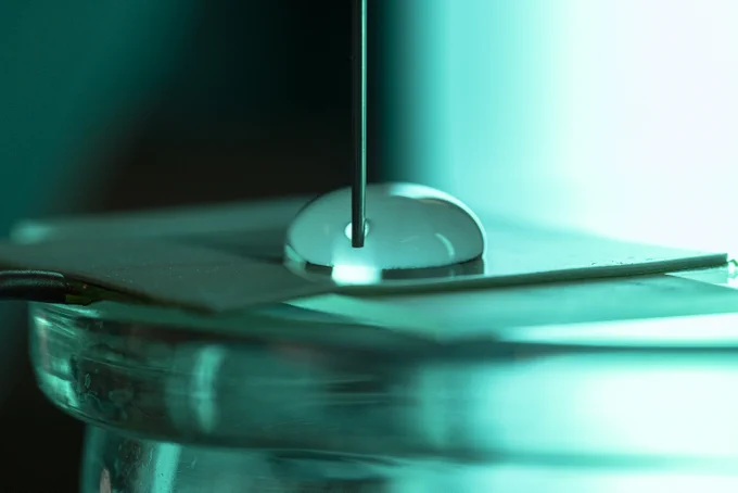 A close up of a drop of water on a plastic surface with a syringe going through the droplet. A turquoise light is used. 