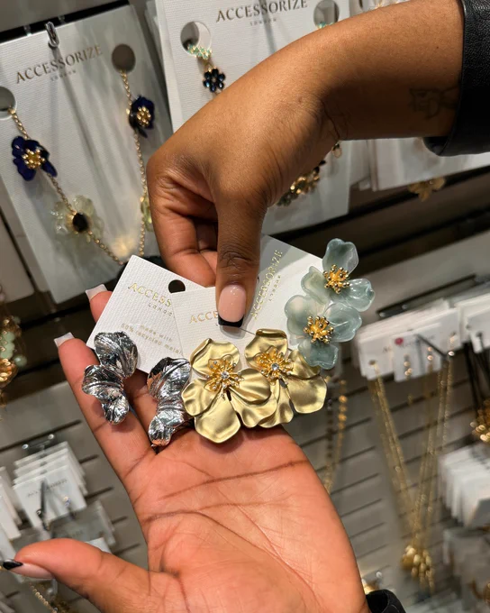 A person's hand holding three floral earrings from Accessorize against a display backdrop filled with jewelry.