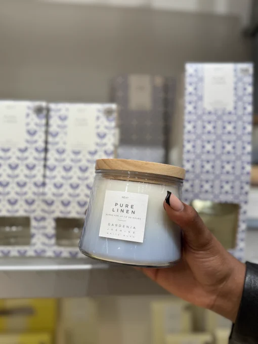 Hand holding a 'Pure Linen Gardenia' scented candle with a wooden lid, in front of a shelf of similarly packaged products.
