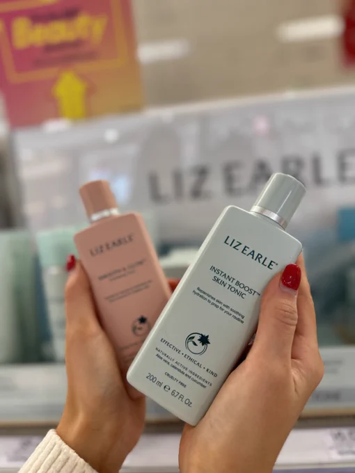 A person holding two Liz Earle skin tonic bottles in a store, one in each hand, with selective focus on the bottle in the foreground.