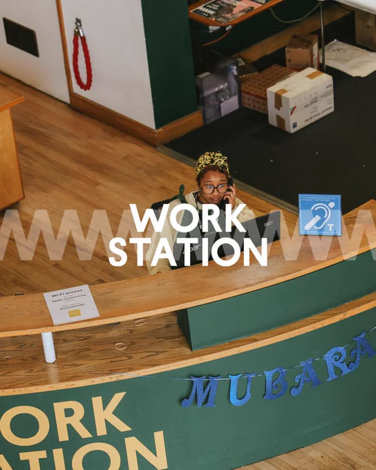 A person wearing glasses and a floral headband peers over a counter with signs labelled "WORK STATION" and "MUBARAK".