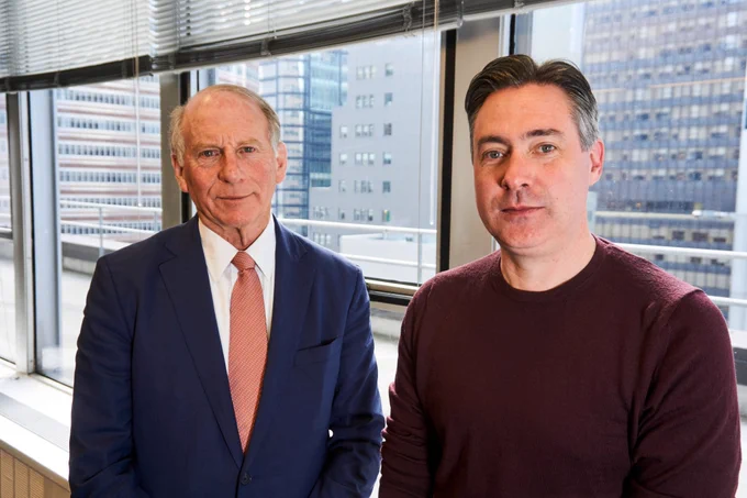 Two men stand beside each other looking at the camera in front of a window