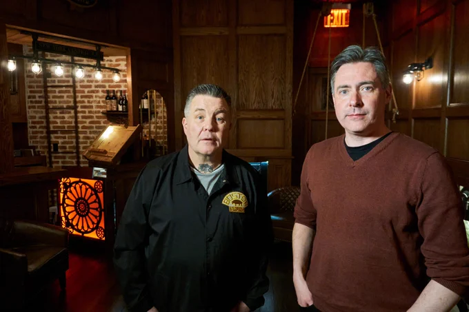 Two men stand next to each other looking at the camera in front of a dark wooden panelled background