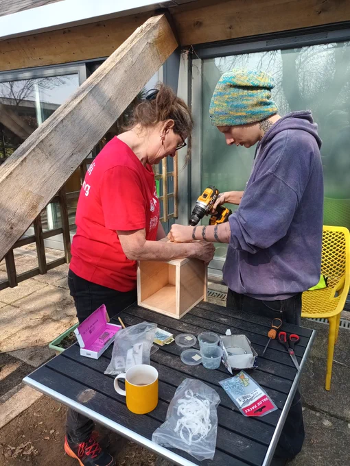 1 individual is holding a wooden composting box while another individual uses a drill to secure the lid