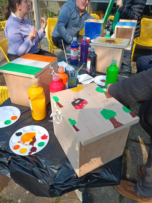People are sitting around a table with 3 painted composting boxes, paints, pallets and paintbrushes in the background

