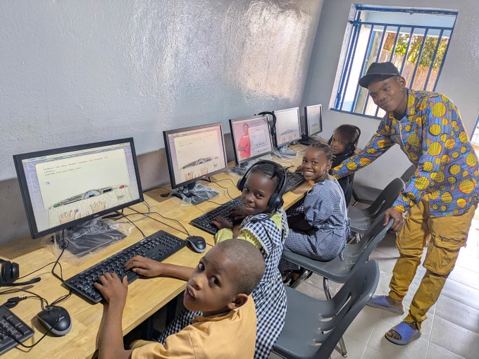 4 young students sit at computers at a long table alongside a wall, with a teacher standing behind them. 